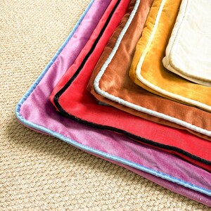 May include: A stack of rectangular velvet pillow covers in various colors including purple, red, brown, gold, and cream. Each cover features a contrasting piped edge in light blue, black, or white, creating a layered and textured appearance.