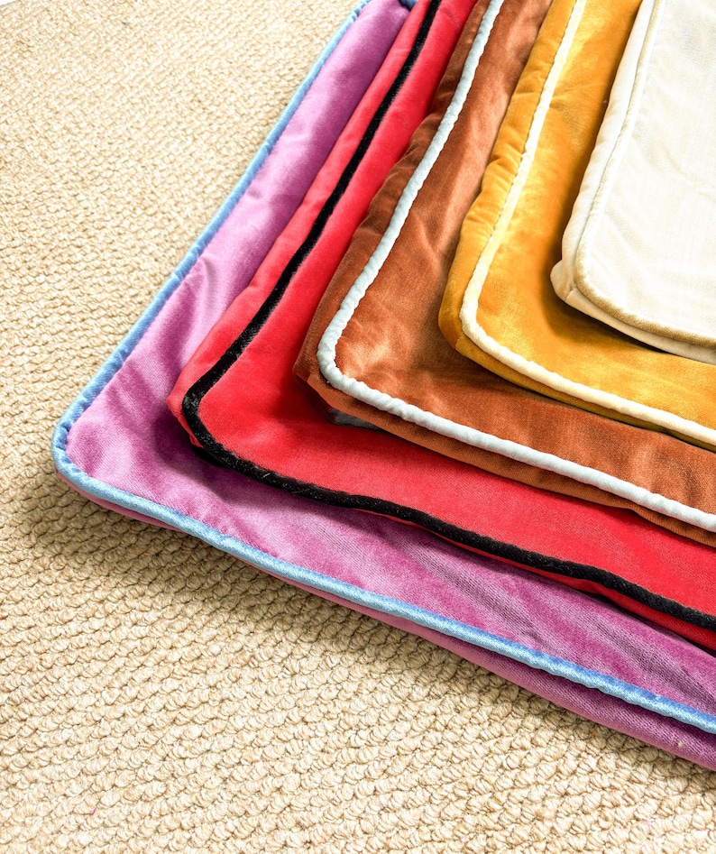 May include: A stack of rectangular velvet pillow covers in various colors including purple, red, brown, gold, and cream. Each cover features a contrasting piped edge in light blue, black, or white, creating a layered and textured appearance.