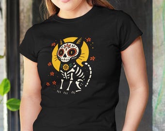May include: Black t-shirt featuring a Day of the Dead cat design. The cat has a skeleton body with a decorated skull, a yellow halo, and orange and yellow floral accents. The design is centered on the shirt.