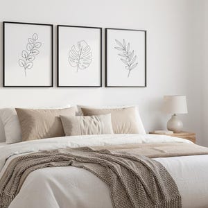Puede incluir: Tres estampados bot&aacute;nicos enmarcados con marcos negros cuelgan sobre una cama. Los estampados presentan dibujos lineales minimalistas de hojas. La cama est&aacute; hecha con s&aacute;banas blancas, almohadas beige y una manta marr&oacute;n. Una l&aacute;mpara de noche se encuentra en una mesita de noche de madera.