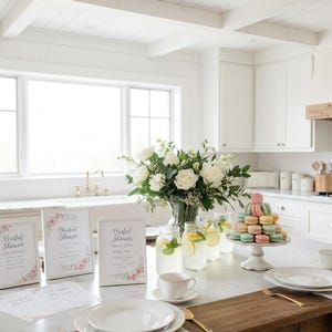 May include: A bright, airy kitchen scene with a marble countertop set for a bridal shower. Features include white roses in a vase, macarons on a stand, lemonade, and framed signs with floral accents.