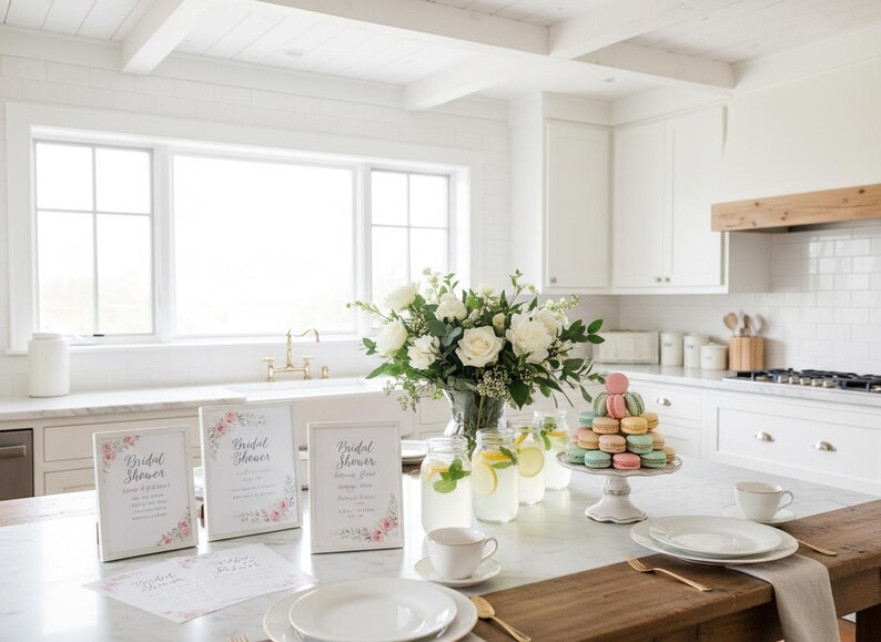 May include: A bright, airy kitchen scene with a marble countertop set for a bridal shower. Features include white roses in a vase, macarons on a stand, lemonade, and framed signs with floral accents.