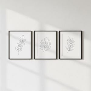 Puede incluir: Tres l&aacute;minas bot&aacute;nicas enmarcadas. Cada l&aacute;mina enmarcada en negro presenta un dibujo lineal minimalista de una planta diferente: una ramita de hojas, una hoja de monstera y una rama de olivo. Las l&aacute;minas se exhiben contra una pared blanca.