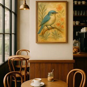 May include: A framed watercolour painting of a bluebird on a branch, with orange flowers and green foliage. The painting hangs on a cream wall above a wooden table with a cup of coffee and a small jar. The scene is set in a cafe.