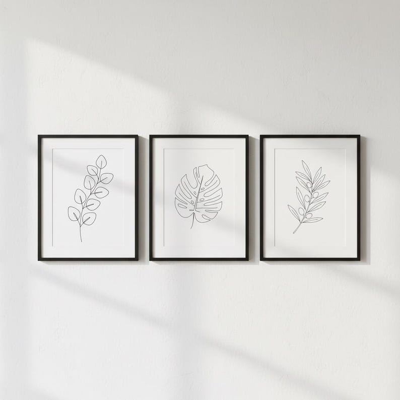 Puede incluir: Tres l&aacute;minas bot&aacute;nicas enmarcadas. Cada l&aacute;mina enmarcada en negro presenta un dibujo lineal minimalista de una planta diferente: una ramita de hojas, una hoja de monstera y una rama de olivo. Las l&aacute;minas se exhiben contra una pared blanca.