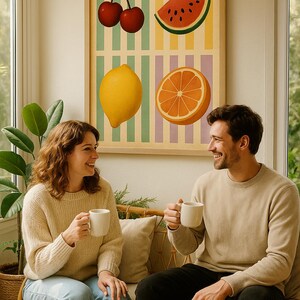 May include: A framed art print featuring illustrations of cherries, watermelon, a lemon, and an orange. The artwork is displayed above a couple seated on a rattan sofa, each holding a white mug. The room has a neutral color palette.