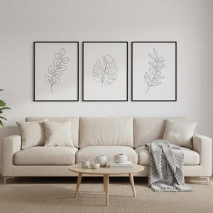 Puede incluir: Tres impresiones art&iacute;sticas bot&aacute;nicas enmarcadas con marcos negros cuelgan sobre un sof&aacute; beige. Las impresiones presentan dibujos lineales de hojas. Una mesa de centro redonda de madera y una planta en maceta completan la decoraci&oacute;n de la sala de estar.