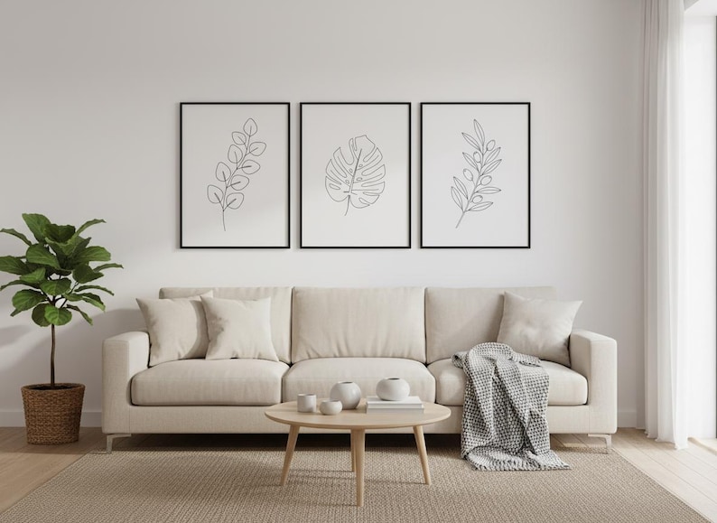 Puede incluir: Tres impresiones art&iacute;sticas bot&aacute;nicas enmarcadas con marcos negros cuelgan sobre un sof&aacute; beige. Las impresiones presentan dibujos lineales de hojas. Una mesa de centro redonda de madera y una planta en maceta completan la decoraci&oacute;n de la sala de estar.