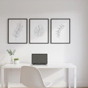 Puede incluir: Tres impresiones bot&aacute;nicas enmarcadas con marcos negros cuelgan sobre un escritorio blanco. Las impresiones presentan dibujos lineales de hojas. Una computadora port&aacute;til est&aacute; sobre el escritorio, junto con una peque&ntilde;a planta y un jarr&oacute;n con vegetaci&oacute;n. Una silla blanca est&aacute; frente al escritorio.