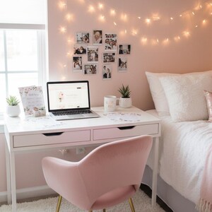 May include: A cozy bedroom scene with a white desk, a pink velvet chair, and a laptop. A bed with white bedding and pink pillows is in the background. String lights and framed photos decorate the pink walls. A small succulent plant sits on the desk.