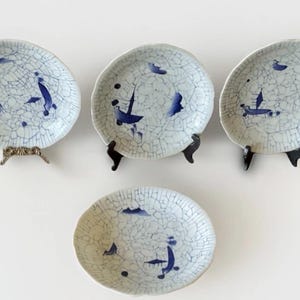 May include: Four decorative ceramic plates with a crackle glaze pattern and blue nautical-themed designs. The plates are displayed on decorative stands. The plates have a white background with blue illustrations of boats and abstract shapes.