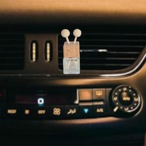 Könnte beinhalten: Ein rechteckiger, klarer Auto-Lufterfrischer aus Glas mit einem Korken aus Holz und zwei weißen Kugeln. Er befindet sich vor einer Lüftungsöffnung des Armaturenbretts eines Autos.