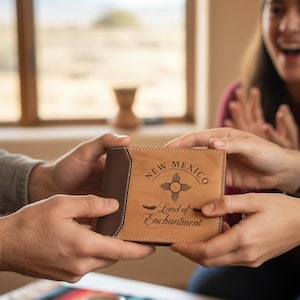 May include: A tan and brown leather wallet with the text "NEW MEXICO" and "Land of Enchantment" is held by two sets of hands. The wallet features a stylized sun symbol. The background is blurred, with a person smiling.