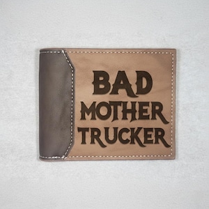 May include: A bi-fold wallet with a brown and tan color scheme. The wallet features the phrase "BAD MOTHER TRUCKER" in dark brown lettering. The left side is a darker brown, with white stitching.