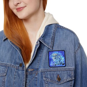 Könnte beinhalten: Ein quadratischer Aufnäher mit einem weißen Tiger mit blauen Akzenten auf einem dunkelblauen Hintergrund. Der Aufnäher ist auf eine Jeansjacke genäht. Der Tiger brüllt, das Maul geöffnet, und ist von einem wolkenartigen Design umgeben.