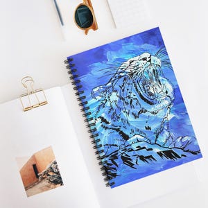 Könnte beinhalten: Spiralgebundenes Notizbuch mit einer lebendigen blau-weißen Illustration eines brüllenden Tigers. Der Tiger ist vor einem strukturierten blauen Hintergrund dargestellt. Das Notizbuch liegt offen auf einer weißen Oberfläche, mit einer goldenen Büroklammer und Sonnenbrille.