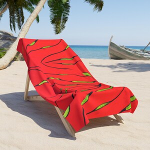 Könnte beinhalten: Ein rotes Strandtuch mit einem schwarzen und grünen Blumendesign ist über einem hölzernen Strandstuhl drapiert. Das Handtuch liegt an einem Sandstrand mit einem Boot im Hintergrund.