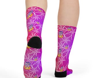Puede incluir: Un par de calcetines con un degradado vibrante en rosa y morado con un patrón de telarañas blancas. Los calcetines están decorados con pequeñas ilustraciones de un gato con sombrero de bruja sentado dentro de una calabaza naranja brillante.