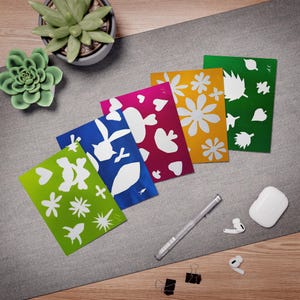Könnte beinhalten: Eine Sammlung farbenfroher Grußkarten mit weißen Blumen- und Tier-Ausschnitt-Designs. Die Karten sind auf einer grauen Oberfläche mit einem Stift, Ohrhörern und einer kleinen Sukkulentenpflanze angeordnet.