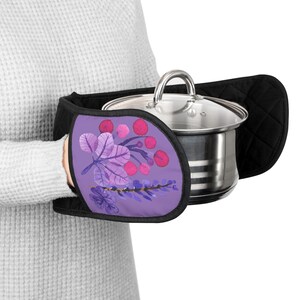 Könnte beinhalten: Schwarze Topflappen mit einem lila Blumendesign, die einen Edelstahltopf mit Glasdeckel halten. Das Blumendesign zeigt lila Blätter und rosa Beeren. Der Topf hat einen silbernen Griff.