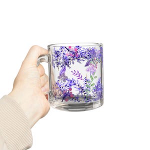 Könnte beinhalten: Transparente Glas-Tasse mit einem floralen Design aus lila, rosa und grünen Blättern und Blumen im Aquarellstil. Die Tasse hat einen Henkel und wird von einer Hand gehalten.