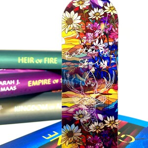 May include: A clear acrylic bookmark with a stained glass design featuring a deer, flowers, and a mountain landscape. The text "Rattle the Stars" is written in white script on the bookmark.