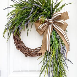May include: A decorative wreath featuring a circular grapevine base adorned with green and purple faux lavender and greenery. A large burlap bow with white and brown stripes adds a rustic touch. The wreath is displayed on a white door.
