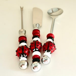May include: A set of three silver serving utensils with ladybug-themed handles. The handles are made of red, black and white glass beads. The utensils include a butter knife, a small spoon and a cocktail fork.