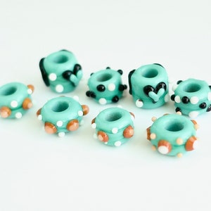 May include: Eight turquoise glass beads with different patterns. Some beads have white and black polka dots, others have brown and white dots, and one has a black heart.