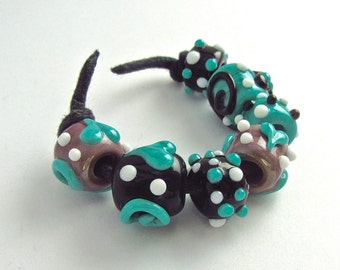 Peut inclure: Un bracelet en cordon noir avec sept perles en verre colorées. Les perles sont noires, blanches, turquoise et brunes avec des pois blancs.