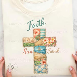 May include: Cream-colored t-shirt featuring a cross design with the words "Faith", "Sun", and "Sand". The cross is composed of patches with floral, seashell, and wave patterns. The design includes teal and gold lettering.