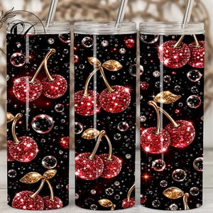 May include: A set of three stainless steel tumblers with a black background. Each tumbler features a pattern of sparkling red cherries with gold stems and leaves, surrounded by bubbles and crystals. The tumblers have silver straws.