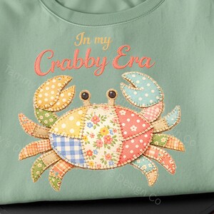 May include: A light green sweatshirt featuring a colorful patchwork crab design. The crab has patterned claws and body segments with floral, polka dot, and gingham patterns. The text "In my Crabby Era" is arched above the crab.