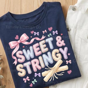 May include: Navy blue t-shirt featuring the words "SWEET & STRINGY" in pastel colours, decorated with bows, hearts, and a pink ribbon. A white denim jacket is partially visible in the background.