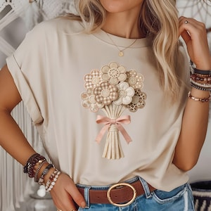 Puede incluir: Camiseta beige con un diseño de ramo de flores de tela. El ramo presenta flores crema, beige y marrón estampadas, atadas con una cinta rosa. La camiseta tiene mangas cortas y un corte holgado. El diseño está centrado en el pecho.