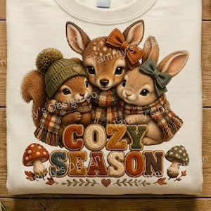 May include: A cream-colored t-shirt featuring a graphic of a squirrel, a fawn, and a rabbit wearing plaid scarves. The squirrel wears a green pom-pom hat, the fawn has a brown bow, and the rabbit has a green bow. Below them is the text COZY SEASON.