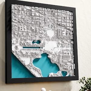 May include: A framed, three-dimensional white map of a city, possibly Washington D.C., with a teal-colored water feature. The map is encased in a black frame and mounted on a wall. The map details buildings, parks, and waterways.