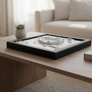 Può includere: Un modello 3D bianco di uno stadio in una cornice nera su un tavolino di legno. Un piccolo vaso con una succulenta e un libro sono anche sul tavolo. Lo sfondo include un divano bianco e una finestra. Il tavolo misura circa 120 cm di lunghezza.