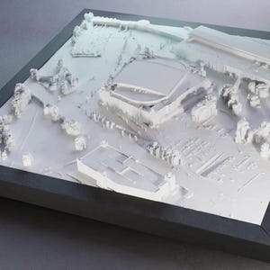 May include: A white 3D architectural model of a building complex, set within a black frame. The model showcases detailed structures, including buildings, trees, and pathways, creating a miniature cityscape. The overall design is minimalist and modern.