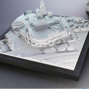 Könnte beinhalten: Ein weißes, 3D-gedrucktes Architekturmodell einer Stadt, gerahmt in einem schwarzen Quadrat. Das Modell zeigt Gebäude, ein Stadion und einen Wasserweg, alles in einem monochromen Stil. Die Details sind komplex und zeigen das Layout der Stadt.