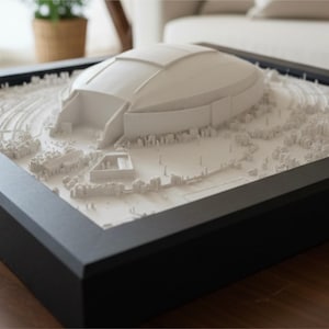 Puede incluir: Un modelo arquitectónico 3D blanco de un gran estadio con techo abovedado, colocado dentro de un marco negro cuadrado. El modelo presenta detalles intrincados del paisaje circundante, incluyendo árboles pequeños, caminos y edificios.