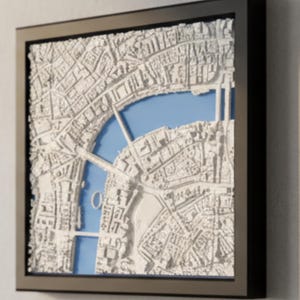 Puede incluir: Un mapa blanco tridimensional enmarcado de una ciudad, posiblemente Londres, con un fondo azul que representa un río. El mapa está encerrado en un marco negro y montado en una pared de color claro. El mapa detalla calles y edificios.
