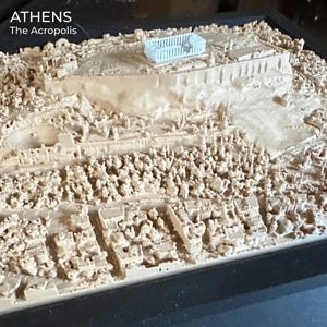 May include: A 3D printed topographical model of Athens featuring the Acropolis. The landscape is rendered in a light beige tone with a distinct pale blue Parthenon structure at the summit. The text ATHENS The Acropolis is printed in white.