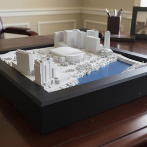 May include: A framed 3D model of a city skyline, featuring white buildings and a blue water feature. The model is set within a black frame and sits on a dark wooden desk. A framed photograph of the city skyline is in the background.