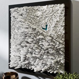 Peut inclure: Une œuvre d'art en relief 3D blanche dans un cadre noir. L'œuvre présente un design texturé et superposé avec une apparence de paysage urbain. Une petite section de couleur teal est visible dans le relief blanc, ajoutant une touche de couleur.