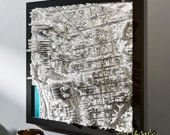 May include: A detailed, three-dimensional white architectural map of a city, framed in black. The map is mounted on a wall, with a glimpse of a teal background. The artwork is a unique home decor piece.