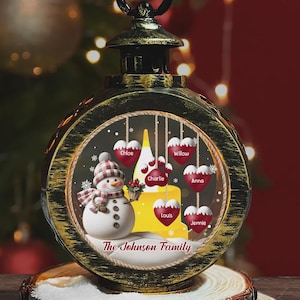 May include: A circular, gold-toned Christmas ornament featuring a snowman and heart-shaped decorations. The snowman has a tartan hat and scarf. The hearts are inscribed with names. The text "The Johnson Family" is at the base.