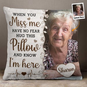 Puede incluir: Una almohada beige con un diseño floral marrón y el texto "When you Miss me Have no fear Hug this Pillow and know I'm here" con un corazón y un gráfico de latido del corazón. La almohada tiene espacio para una foto y un nombre personalizado en forma de corazón.
