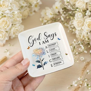 May include: A square, white ceramic trinket dish with gold edging. The dish is inscribed with the words "God Says I AM" and a floral design. Additional text includes "Adorned," "Loved," "Image," "Called," and "Eternal."