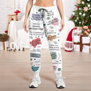 Puede incluir: Pantalones deportivos blancos con cordón negro y varias afirmaciones impresas en un diseño colorido. El texto incluye frases como "I am fucking unstoppable" y "I'm a goddamn ray of sunshine."